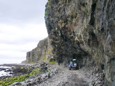 Ein Jeep der unter einen Überhang einen Berg entlang der Küste von Svalvogar fährt